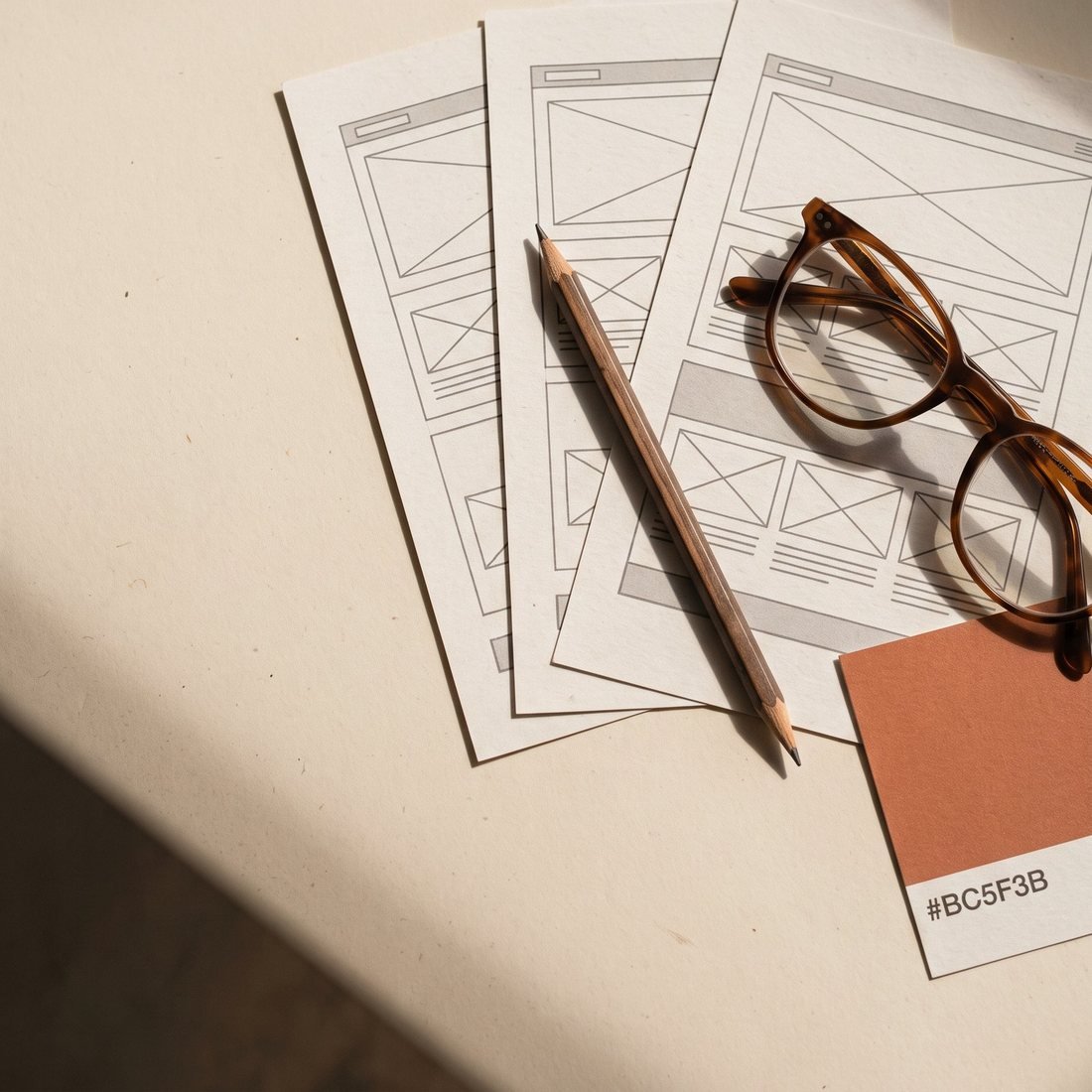 Printed page wireframes fanned out with a pencil, reading glasses, and a clay colour swatch